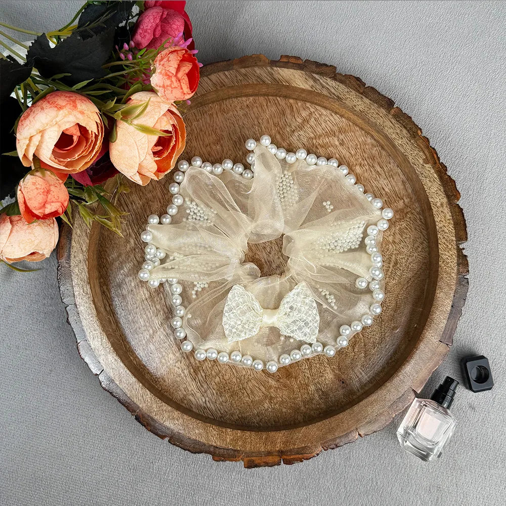 Decorative hair scrunchie with pearls on a wooden surface with flowers and perfume bottle in the background.