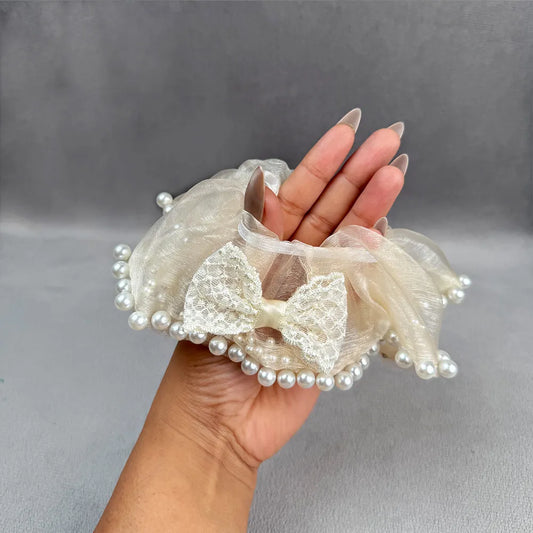 Hand holding a decorative bow scrunchie with pearls against a gray background